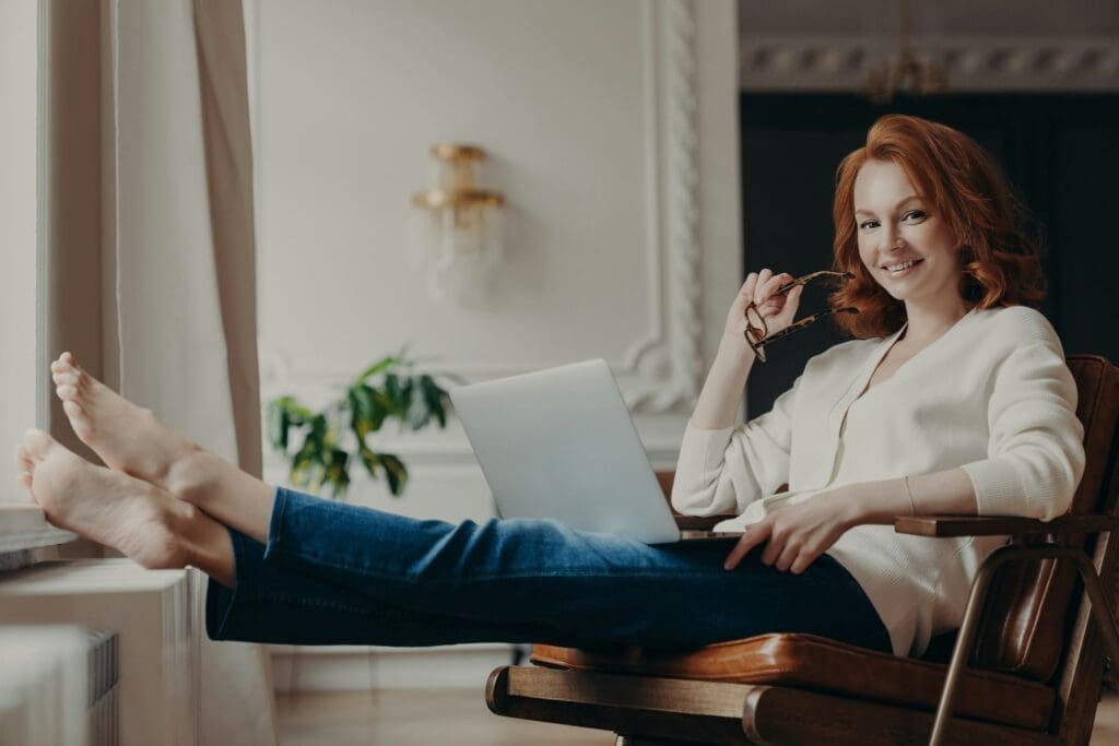 Cheerful successful ginger woman copywriter works from home, keeps laptop on knees